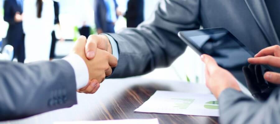 People in business suits shaking hands, with a tablet and documents on a table.