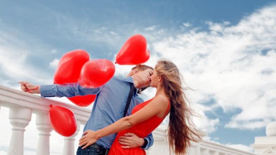 Una coppia si bacia teneramente all'aperto, circondata da palloncini rossi a forma di cuore.